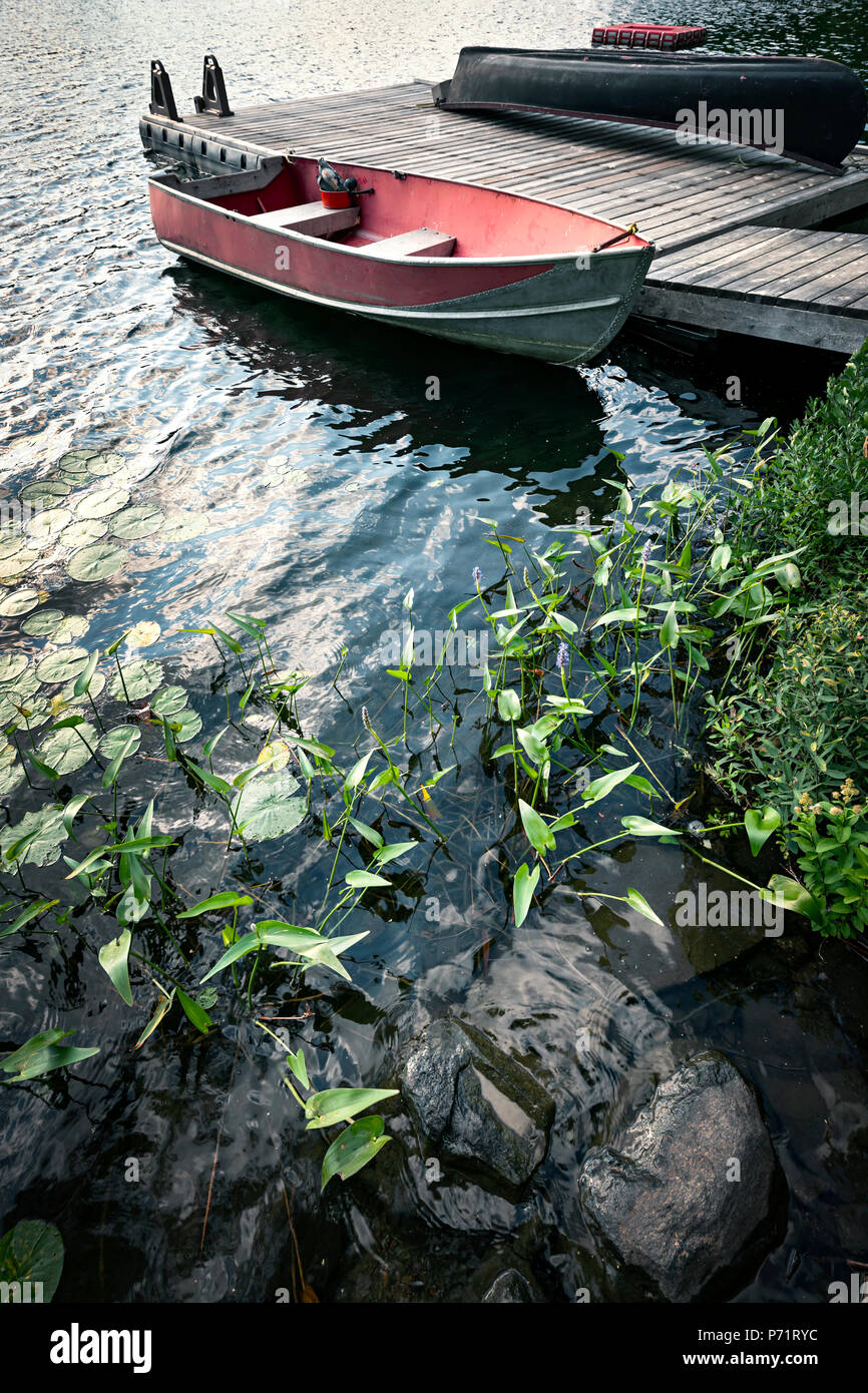 Red boat at wooden dock on lake in cottage country with foreground of rocks and plants Stock Photo