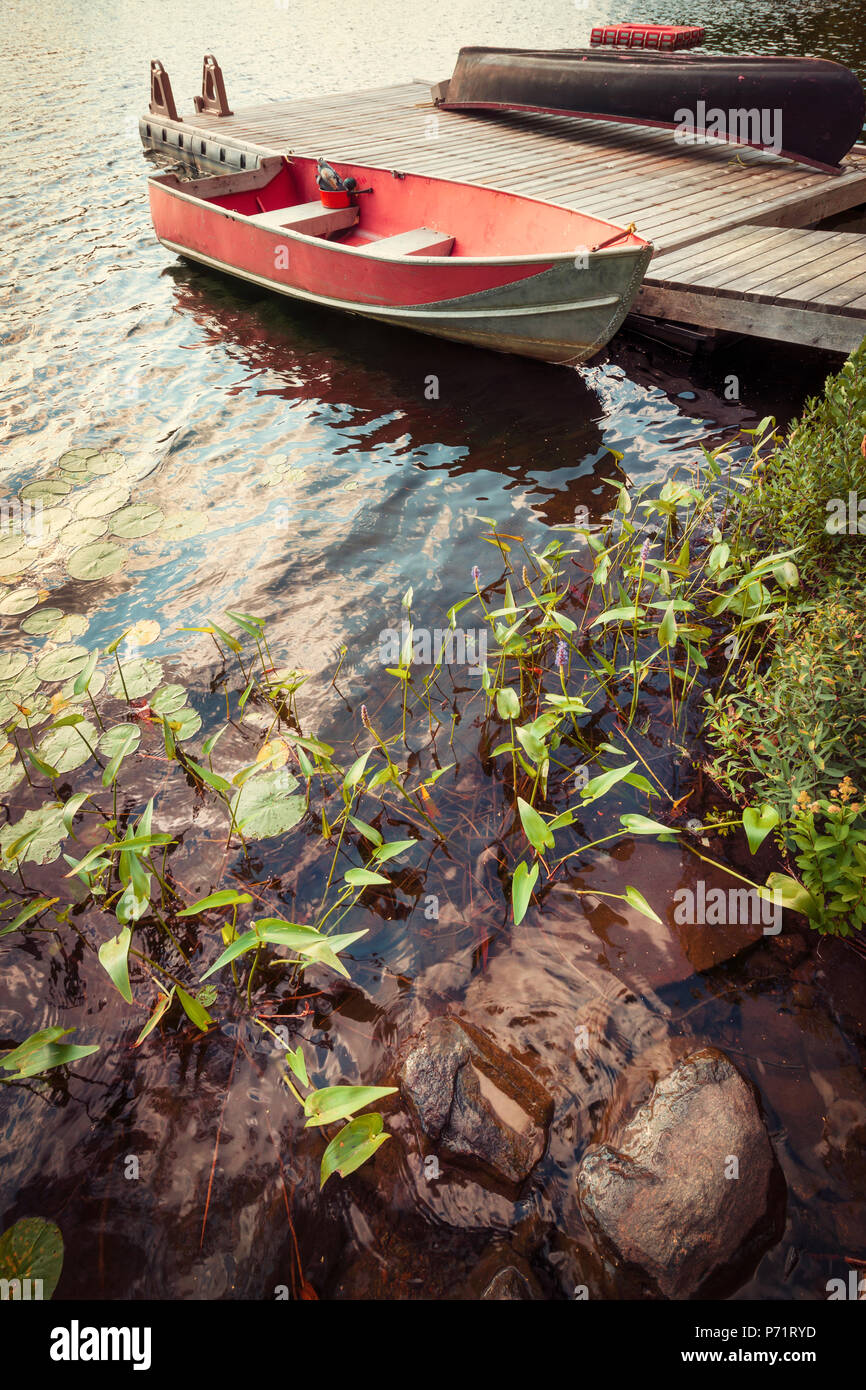 Red boat at wooden dock on lake in cottage country with foreground of rocks and plants Stock Photo