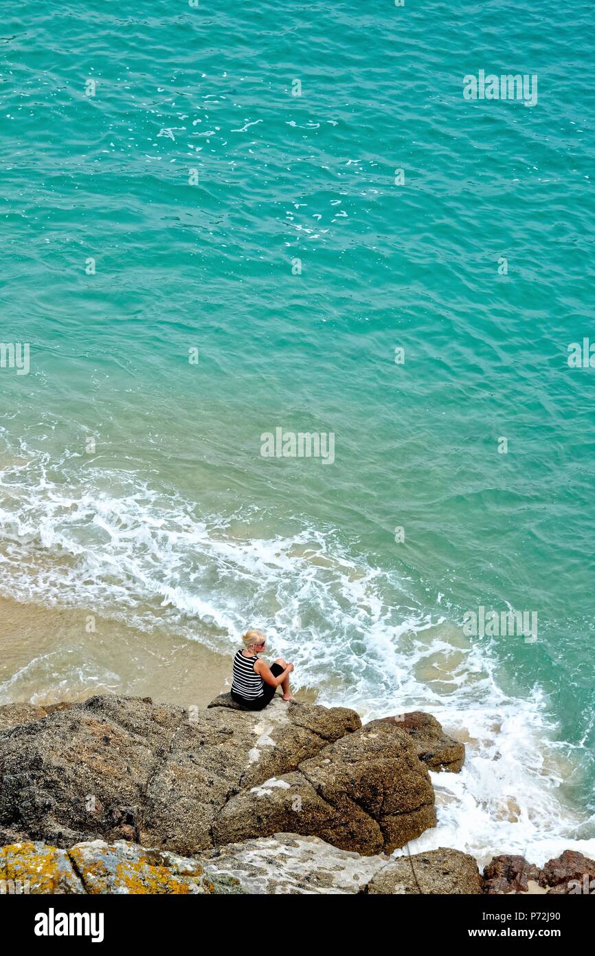 Middle aged blonde woman sitting on rocks surrounded by a Cornish blue sea Porthcurno Cornwall England UK Stock Photo