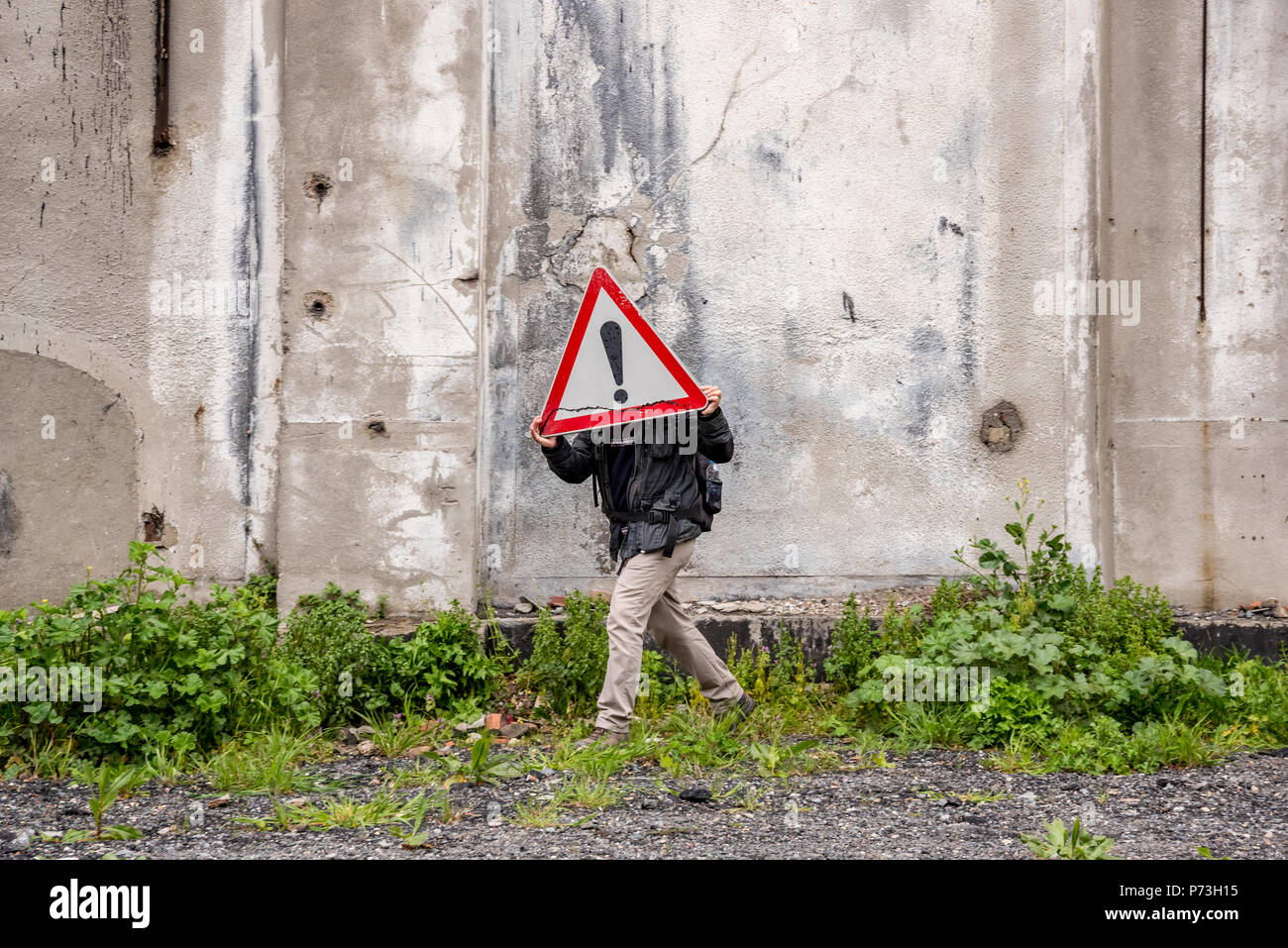 Unidentified man holds a red traffic triangle warning sign in front of his head near a ruined building. Stock Photo