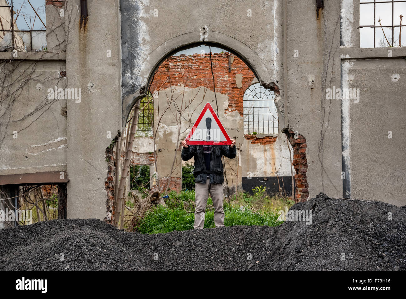 Unidentified man holds a red traffic triangle warning sign in front of his head near a ruined building. Stock Photo