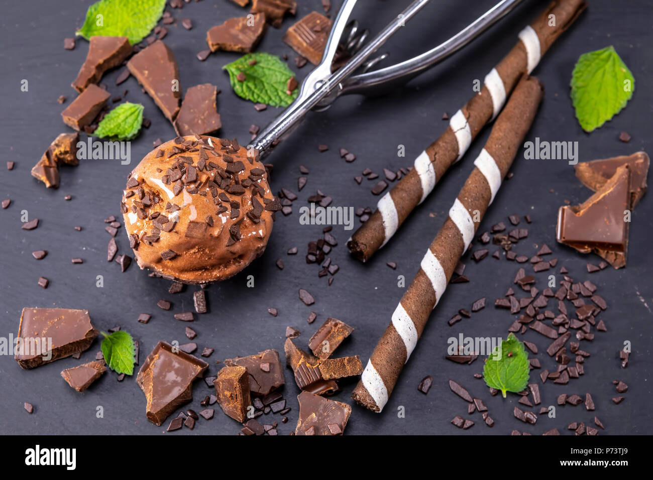 Chocolate ice cream in scoop with wafer sticks and chocolate on a black slate board. Focus on ice cream in scoop. Stock Photo