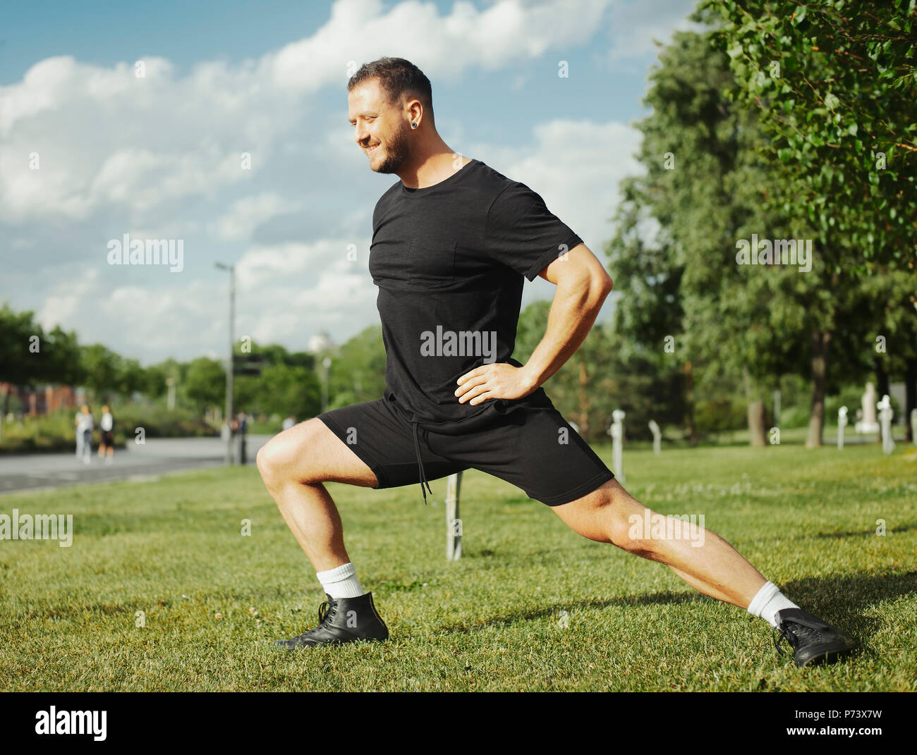 Young attractive man doing lunge outdoor in the park. Side view. Stock Photo