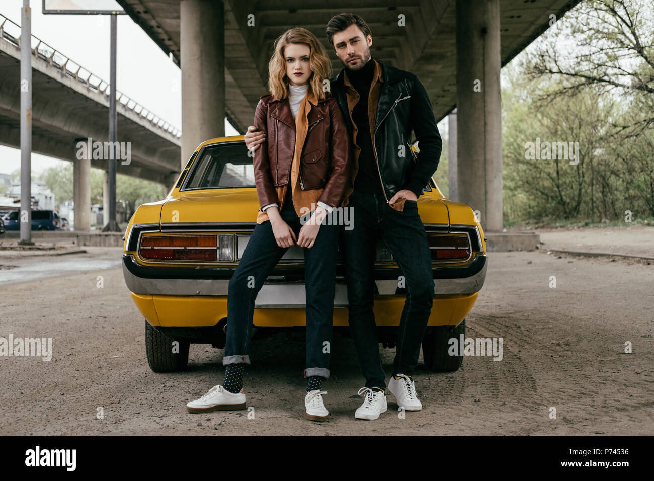 full length view of young couple in leather jackets sitting on yellow old-fashioned car and looking at camera Stock Photo