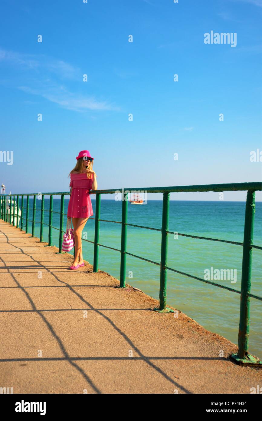 Young girl in pink sundress and with handbag standing on a pier leaning ...
