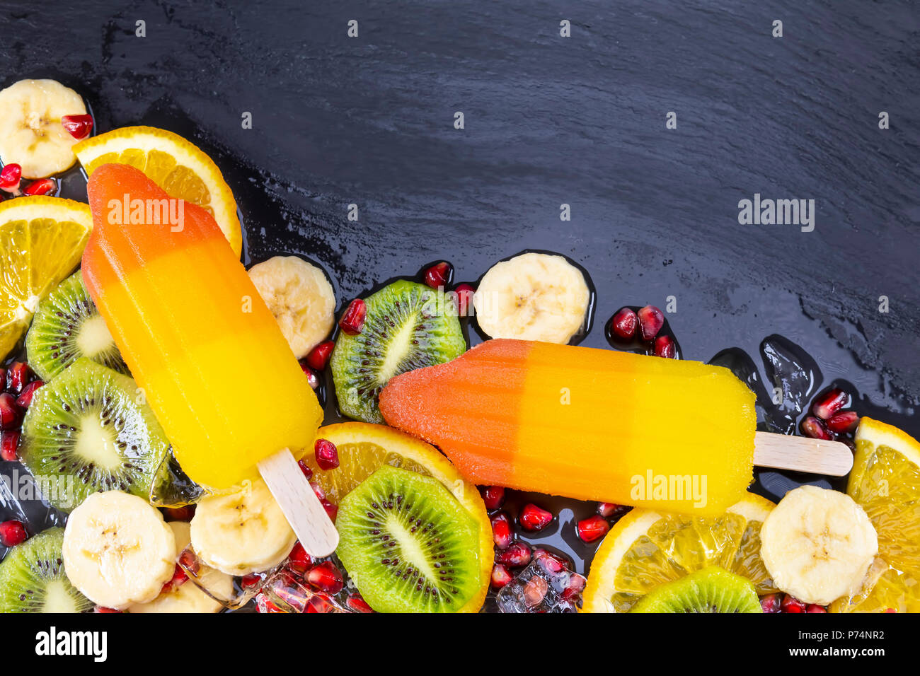 Fruit ice cream on stick with slices fruits on black slate board. Space for text! Focus on Popsicles. Stock Photo