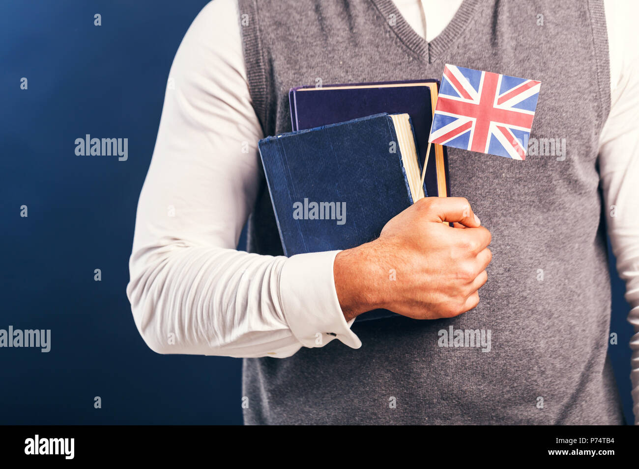 Man wears grey sweater vest holds english books and flag before dark blue studio background
