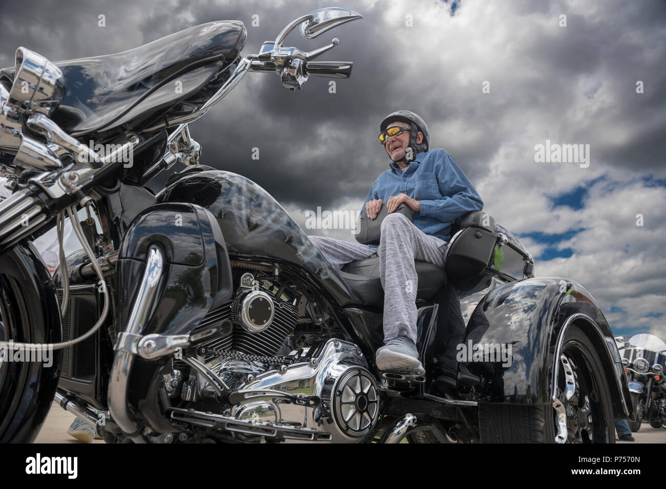 Elderly man smiles broadly as he sits on the back of a powerful three-wheeled motorcycle on a stormy day with dark clouds above him, nearby trees, clo Stock Photo
