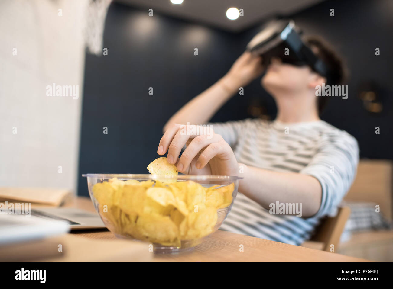 Young male sitting at home reaching for potato chip in bowl while focused on using virtual reality headset. Stock Photo