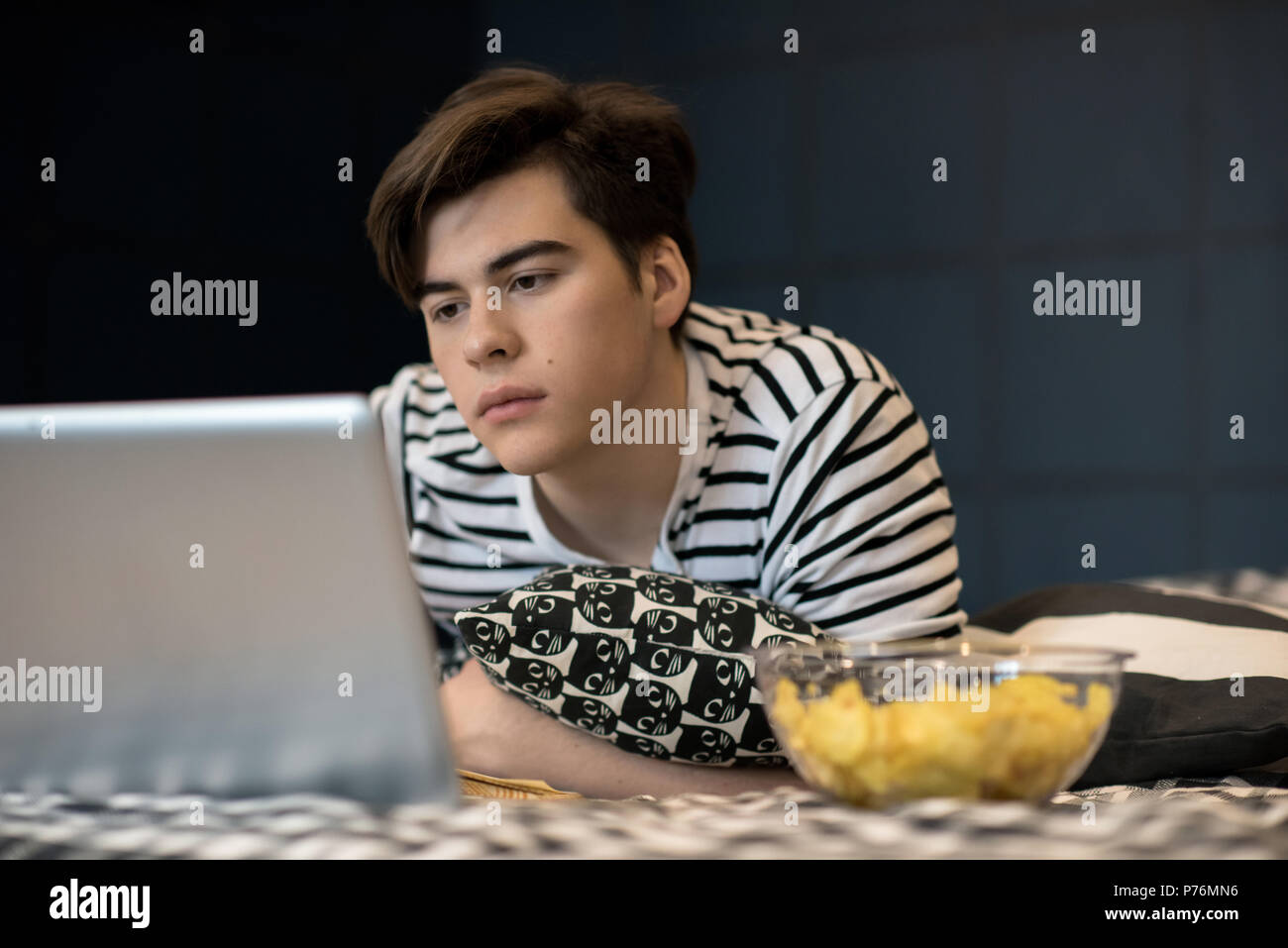 Handsome young male lying on bed with bowl of chips and reading on laptop screen. Stock Photo