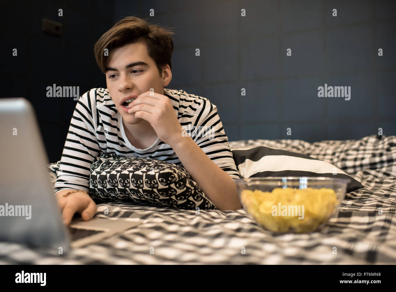 Attractive young male lying on bed and eating chips concentrated on using laptop computer. Stock Photo