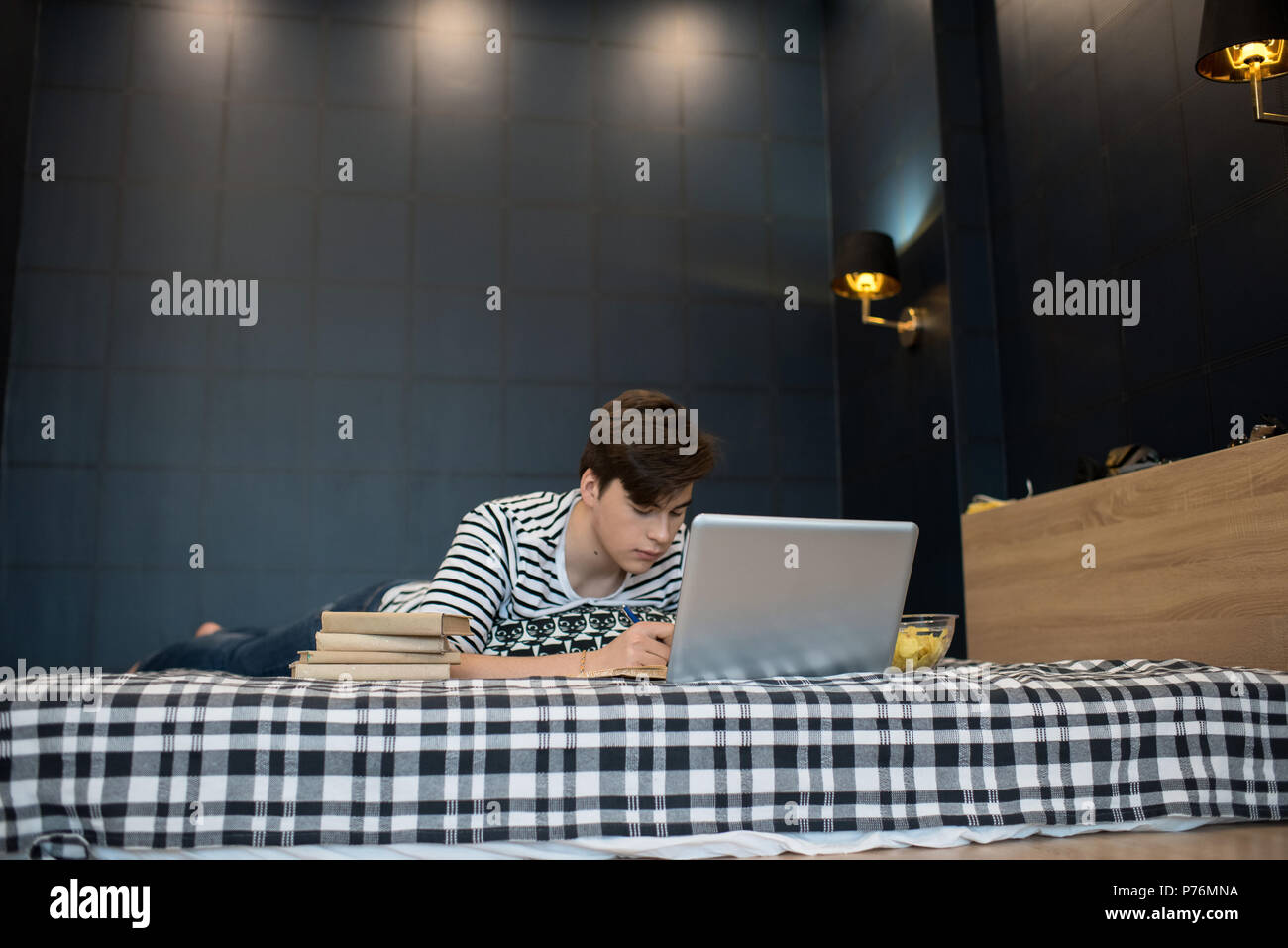 Attractive young male lying on bed with stack of books using laptop computer and taking notes. Stock Photo