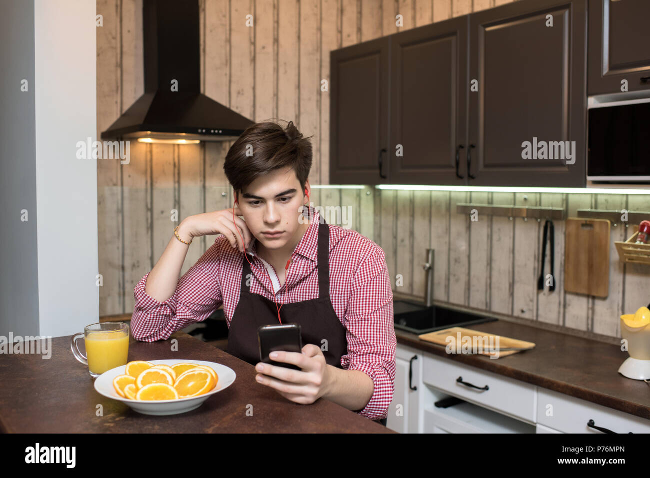 Young man enjoying good music and using smartphone while sitting in cozy kitchen alone. Stock Photo