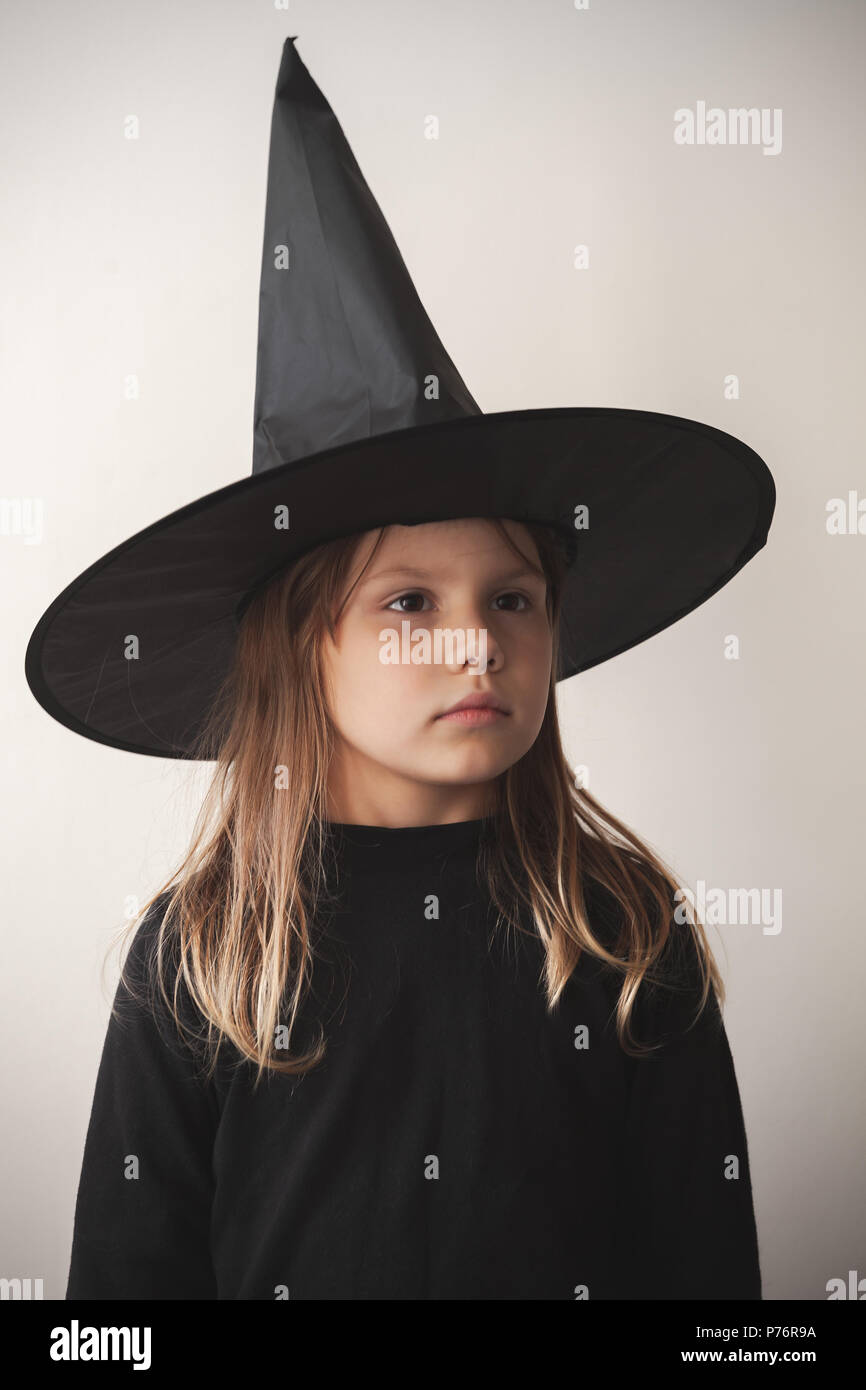 Serious little blond European girl in black witch costume over white wall, close-up studio portrait Stock Photo