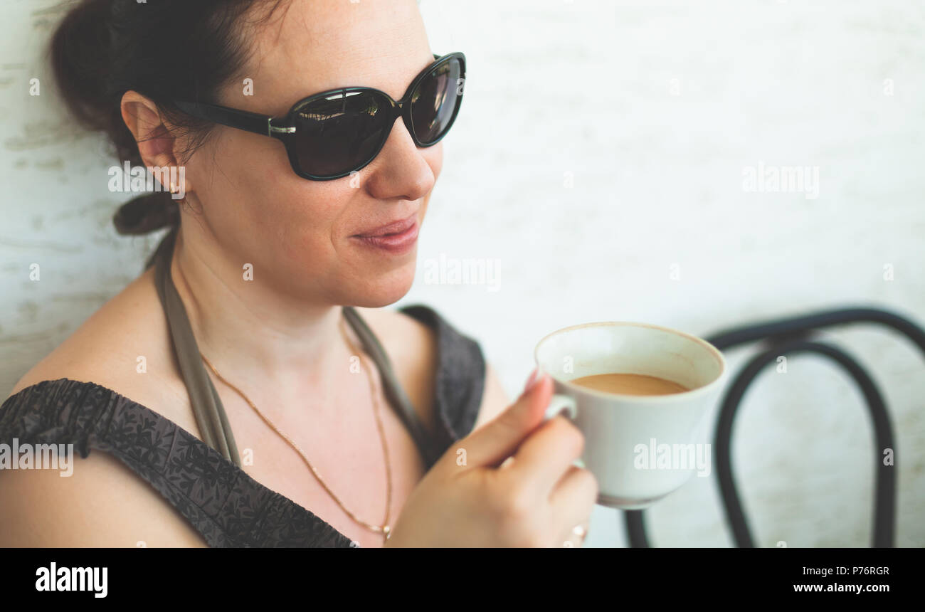 Young adult European woman in dark sunglasses drinks coffee on patio Stock Photo