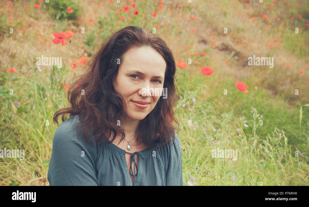 Smiling young adult European woman on a summer meadow with flowering poppies. Vintage toned outdoor portrait Stock Photo