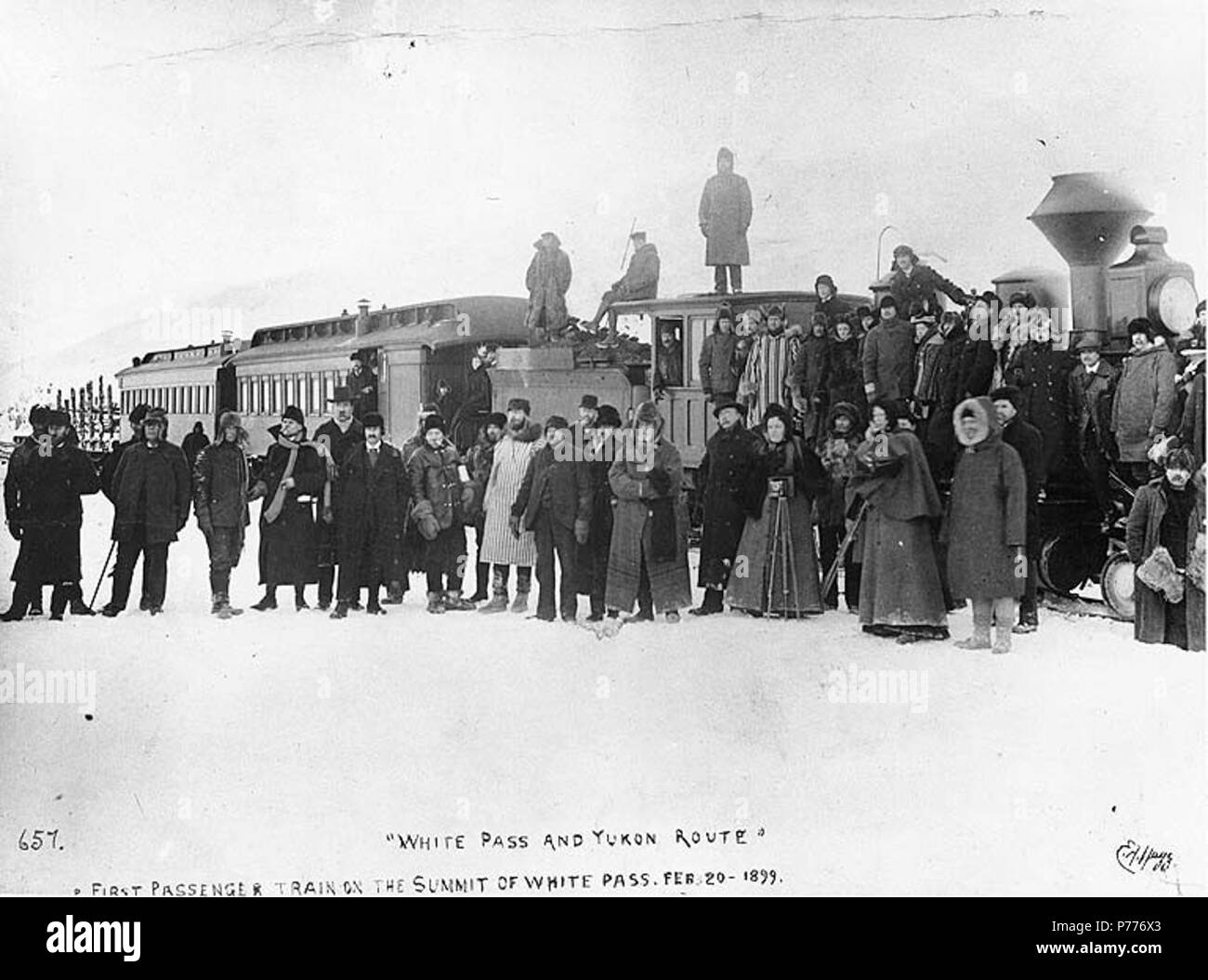 30 men and 2 women dressed in black and white English: First passenger train of the Pass & Yukon Railroad on the summit of White Pass, Alaska, February 20, 1899. English: Shows crowd of women, some with cameras RP_Flexible