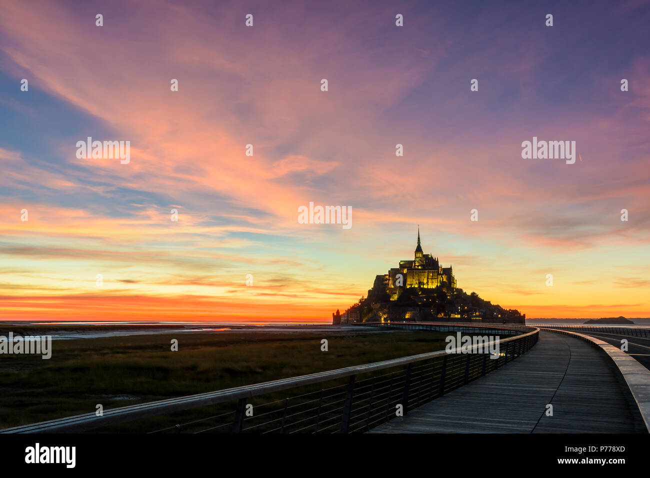 View of the Mont Saint-Michel tidal island in Normandy, France, at nightfall with the wooden jetty in the foreground and purple clouds in the sky. Stock Photo