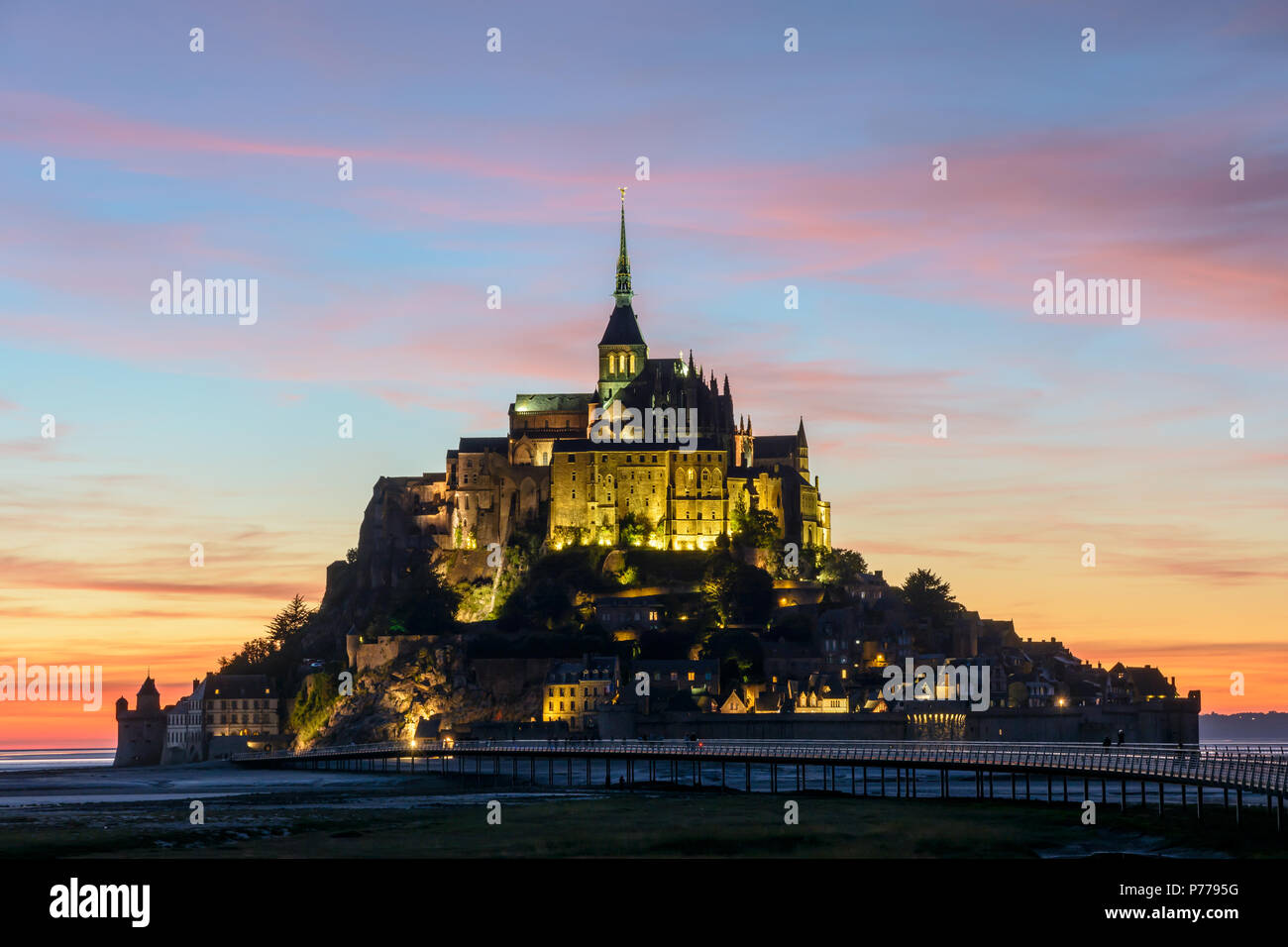 View of the Mont-Saint-Michel tidal island in Normandy, France, illuminated at nightfall with the jetty on stilts and purple clouds in the sky. Stock Photo