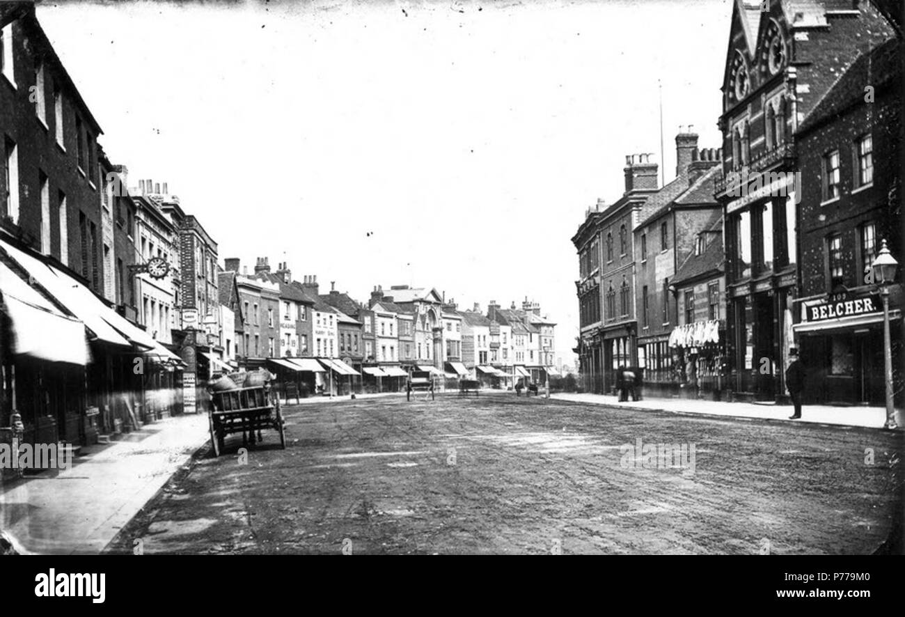English: Broad Street, Reading, looking eastwards, c. 1870. North side: No. 28 (John Player, watch maker, with a clock on a bracket outside the shop); No. 27 (Woolpack Inn); No. 26 (E. Fisher, Cookham Shoe Warehouse); Nos. 15 and 16 (Harry Sims, linen draper); entrance to the covered market and Corn Exchange; No. 6 (John Stow, mineral water manufacturer and wholesale confectioner). South side: No. 113 (James Macaulay, printer and bookseller); No. 112 (Edward Woodman, tailor and woollen draper); Nos. 110 and 111 (Heelas, Sons and Company, drapers); and No. 109 (John Belcher, butcher). 1870-1879 Stock Photo