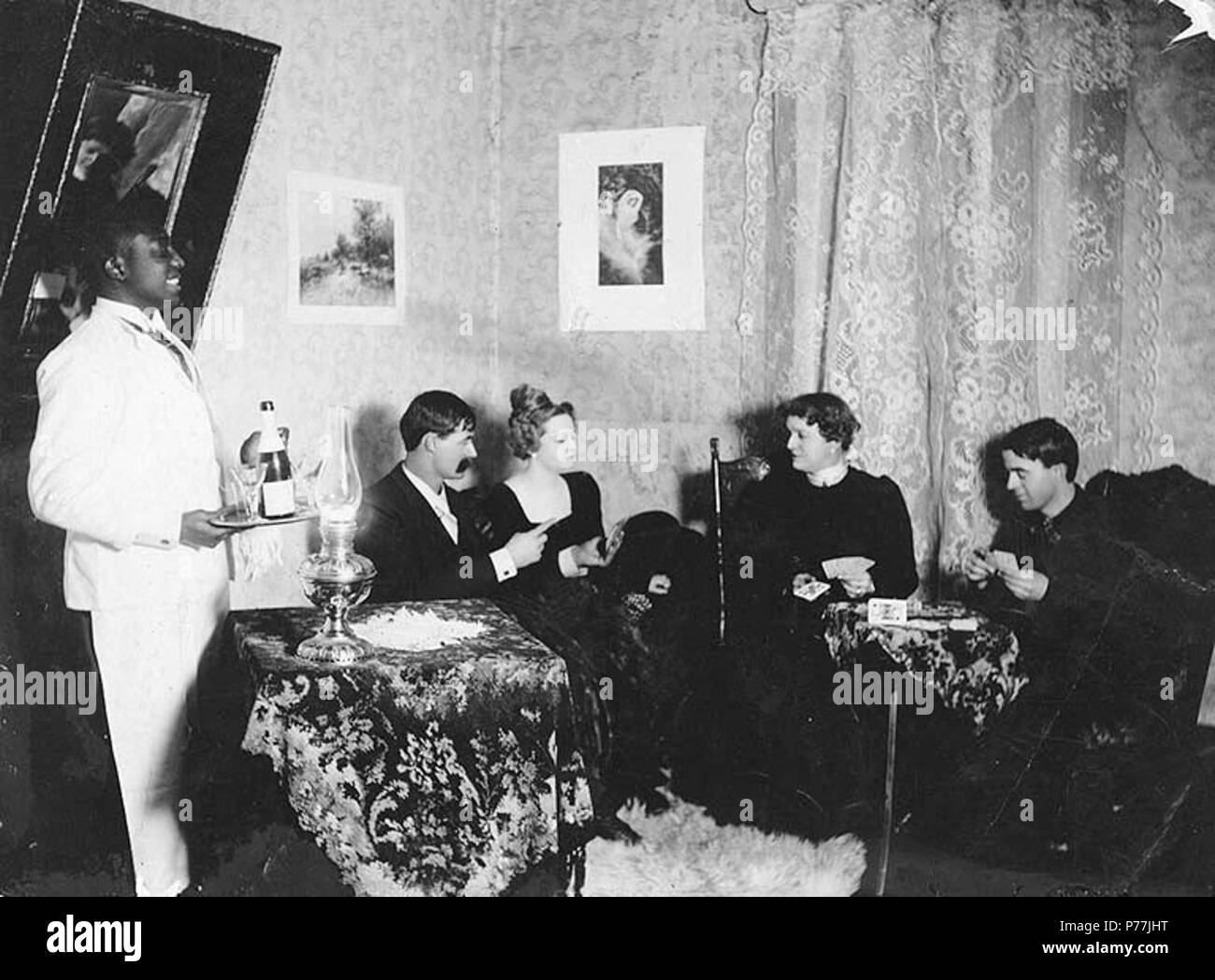 . English: Social scene showing interior of parlor, Dawson, Yukon Territory, ca. 1898. English: Parlor scene with men and woman seated eating, drinking and playing cards. Afro-American man is standing to the left . Caption on image: 'A social call on one of Dawson's fairest. Hegg & Co. Flash Light...Y.T.' Left to right are Black Prince, a prize fighter connected with the Monte Carlo; a man called Jack; Gertie Lovejoy, best known as Diamond Tooth Gertie; Cad Wilson, … ; and Tommy Dolan, brother of comedian Eddie Dolan. (Murray Morgan, One Man's Gold Rush, p. 161) Original image in Hegg Album B, Stock Photo
