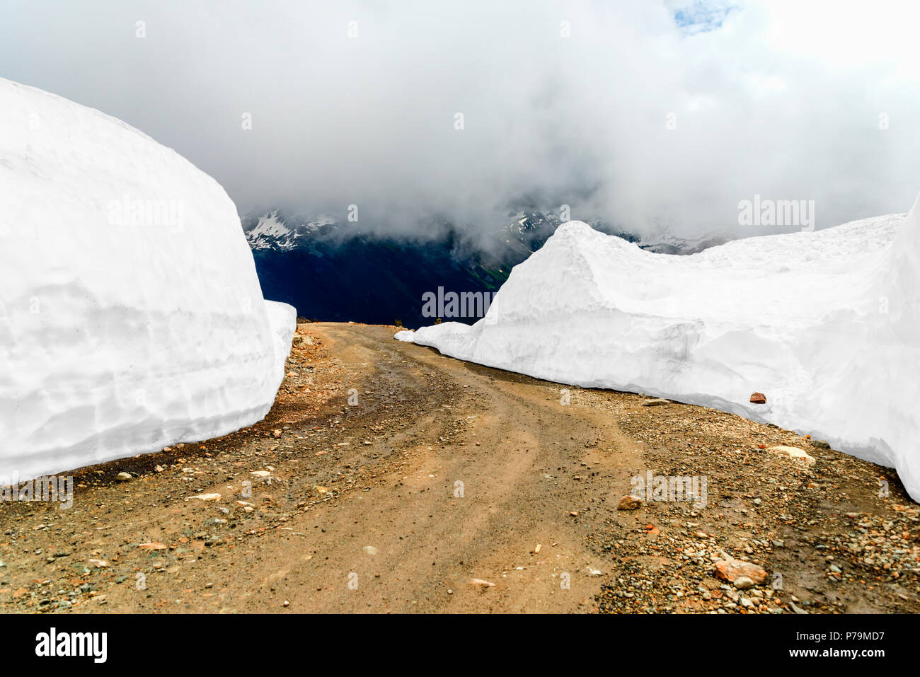 A ground, stony road in the mountains, between two blocks of snow, white mist and mountains in the background Stock Photo