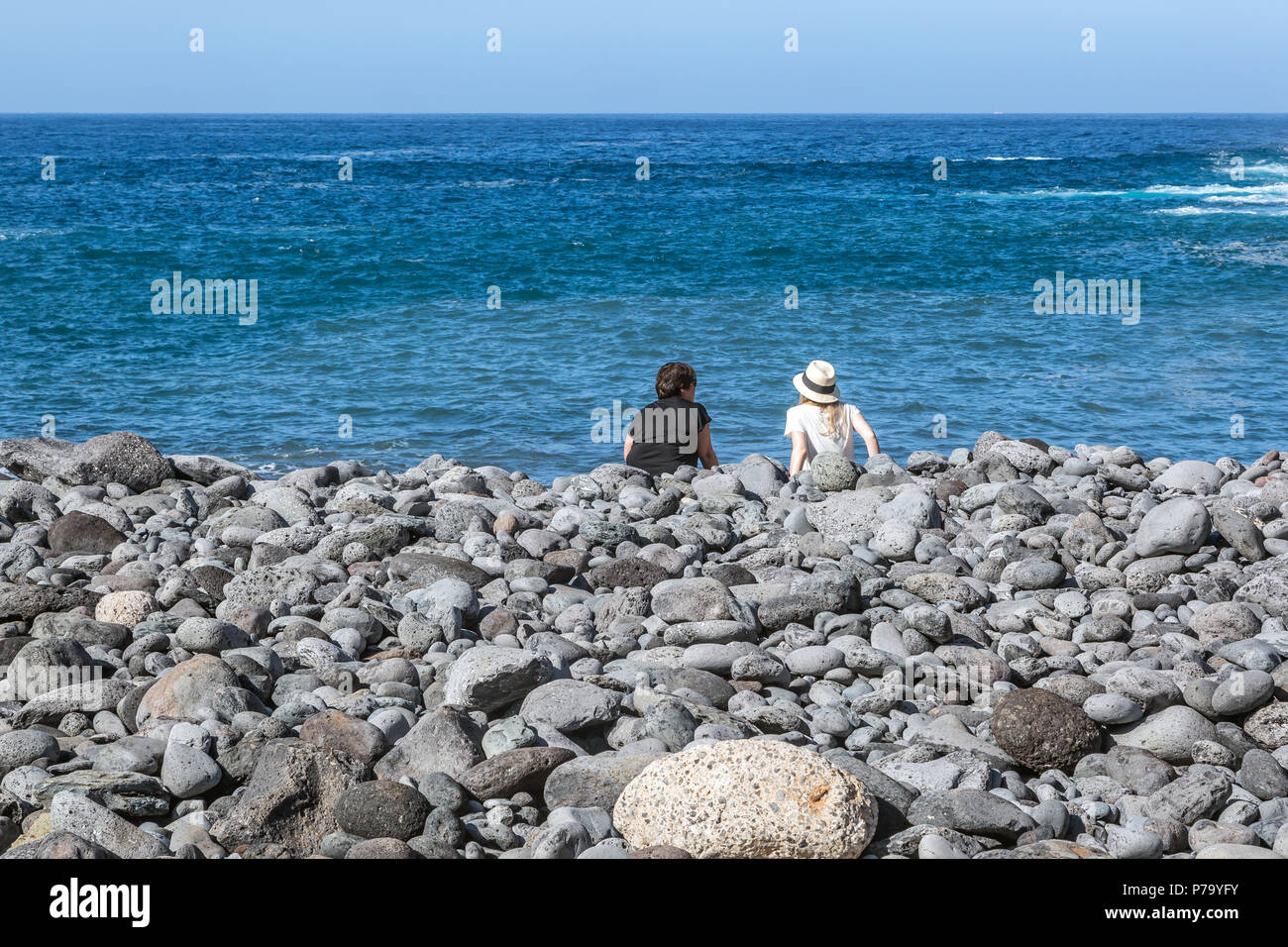 A rear view of a Mother and Daughter sitting on a pebbled beach looking towards the North Atlantic Ocean. Tenerife, Canary Islands. Stock Photo
