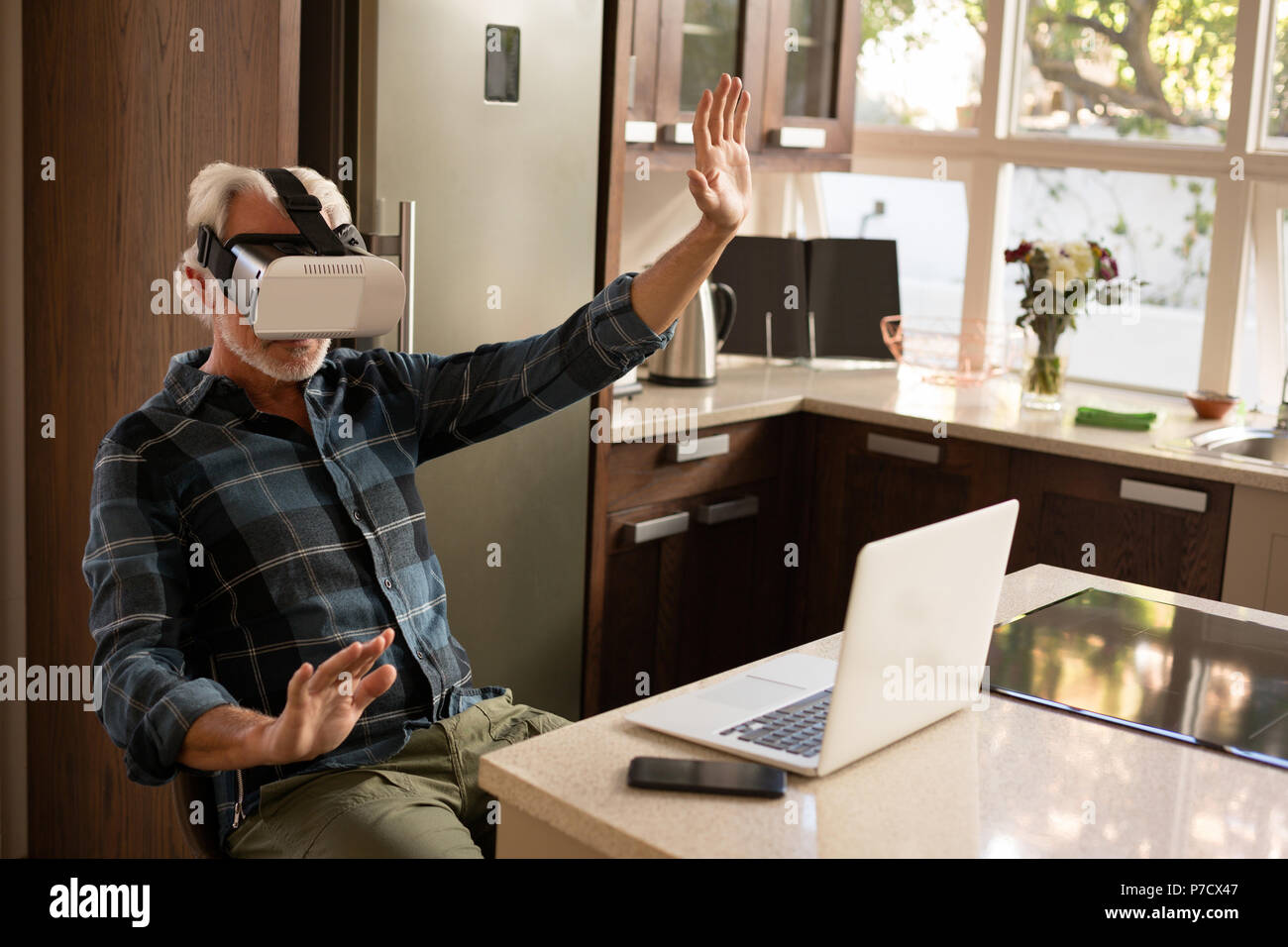 Senior man using virtual reality headset with laptop in kitchen Stock Photo
