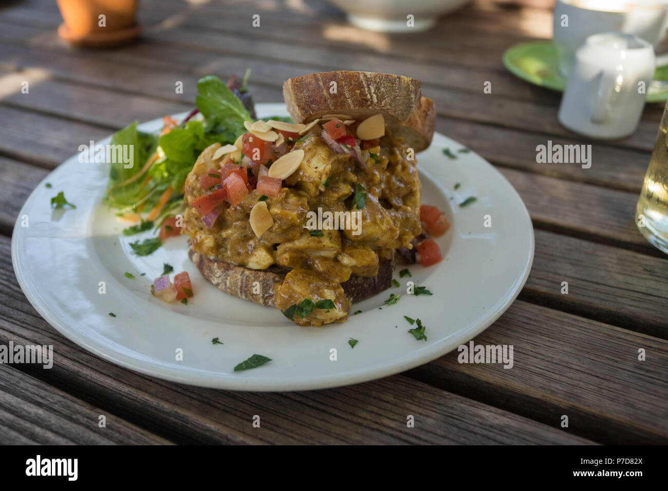 open sandwich filled with grilled chicken in spicy harissa and date dressing on white plate with salad placed on top of wooden slatted table Stock Photo