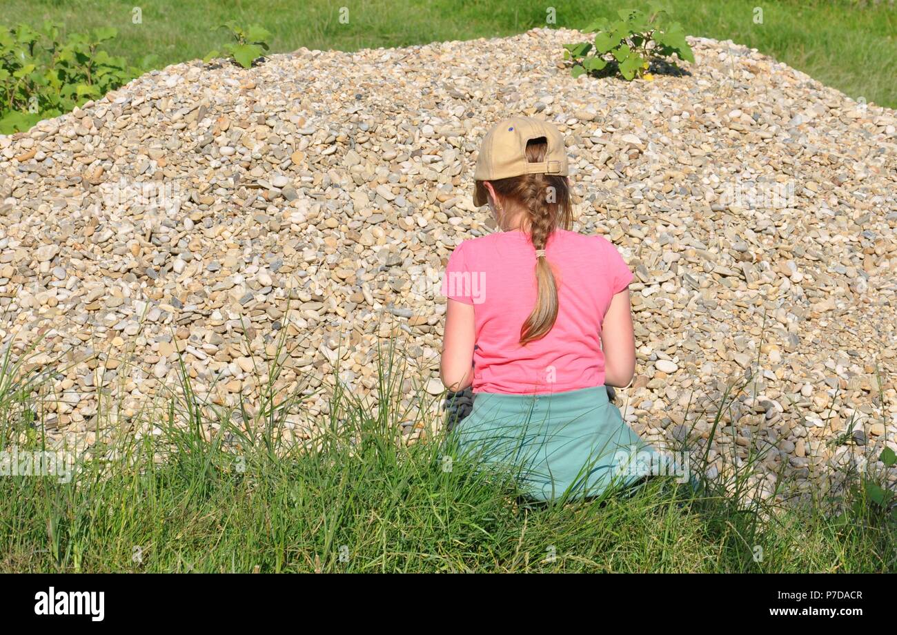 Blonde child girl, (7-9) standing with her back in front of a heap (pile) of stones and playing, copy space Stock Photo