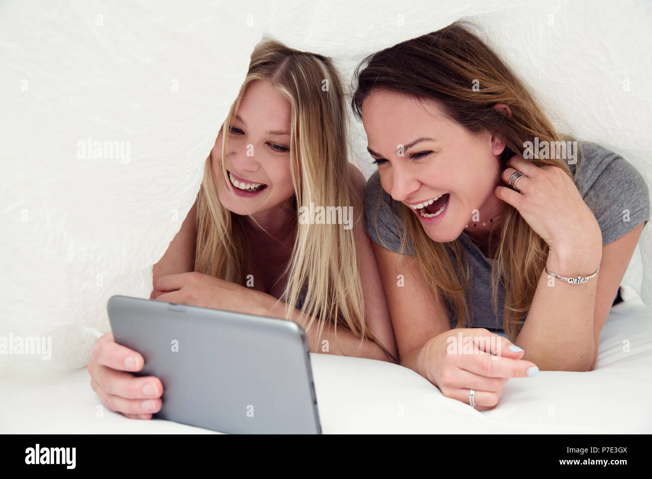 Friends underneath bed sheet using digital tablet smiling Stock Photo