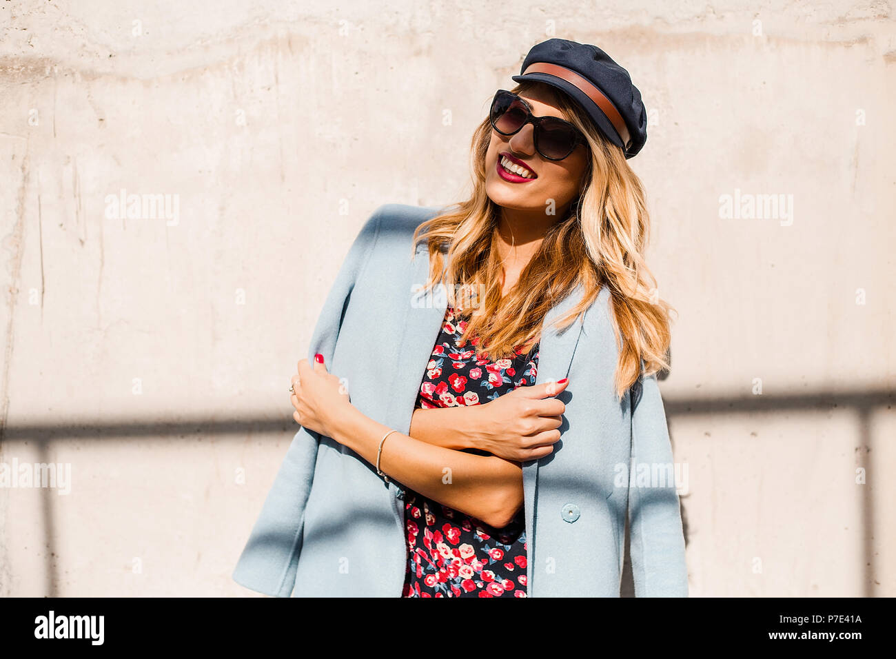 Stylish woman wearing sunglasses and cap in front of wall with arms folded Stock Photo