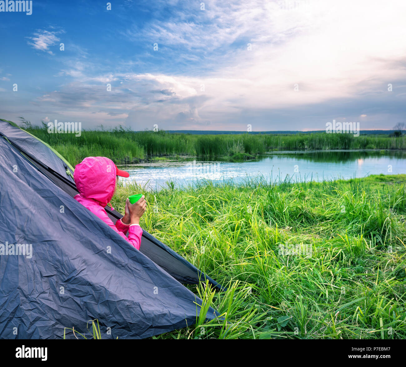 Girl in pink jacket sits at the exit of tent with mug and looks into the distance. Summer landscape. The concept of travel and freedom. Stock Photo