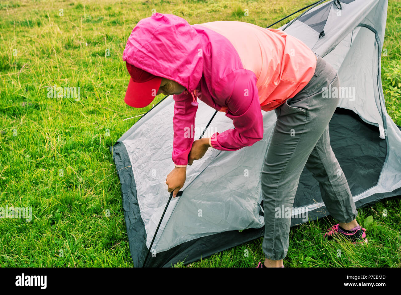 Girl in pink jacket with hood sets tent on a green grass Stock Photo