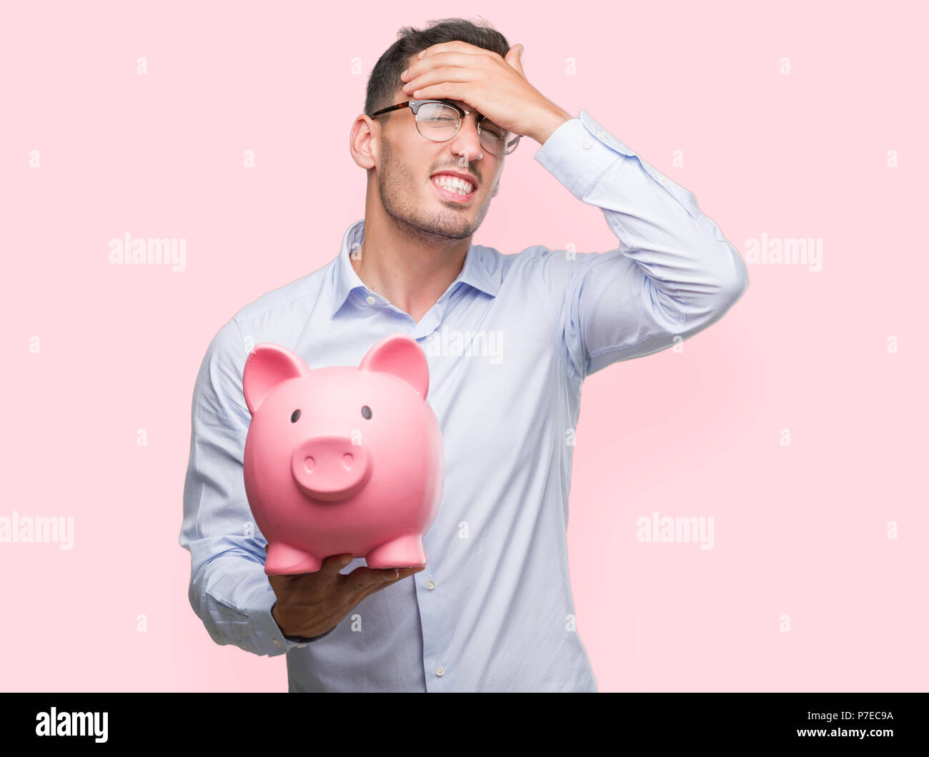 Handsome young man holding a piggy bank stressed with hand on head ...