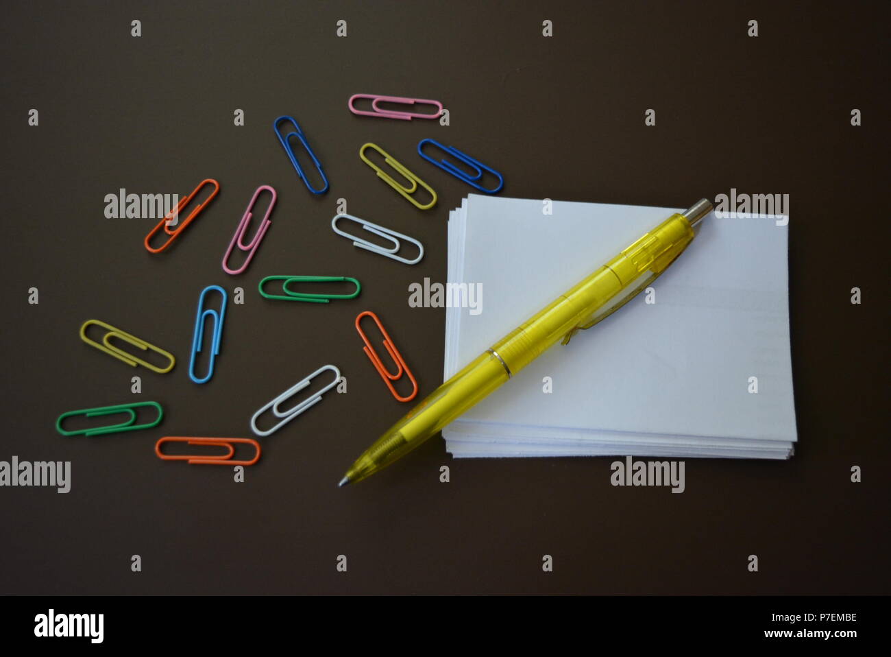 Color paperclips of different colors with sheets of white paper for recording and a yellow pen on a brown matte background. Stock Photo