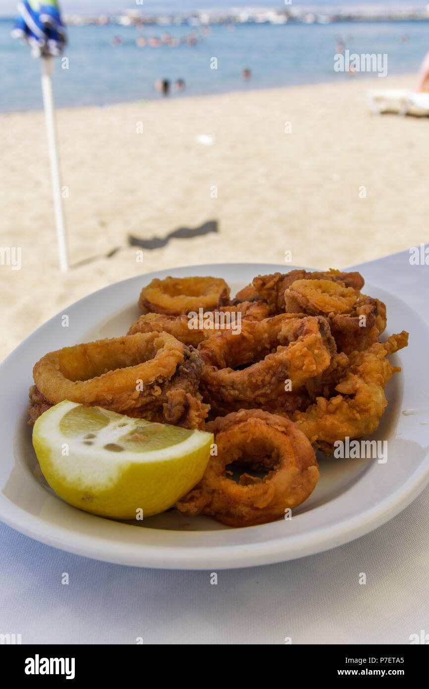 Greek tavern restaurant food. Fried squid in slices served on a white dish with lemon in blurred beach background. Stock Photo