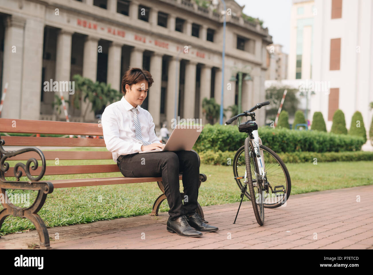 Young handsome businessman with laptop and bicycle sitting on bench in park Stock Photo