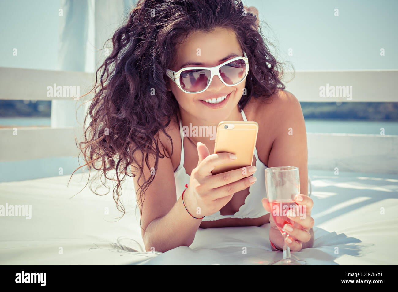 Closeup young beautiful happy, excited smiling woman texting, looking on mobile cell phone drinking champagne outdoors on sun-bed at sea, lake. Positi Stock Photo