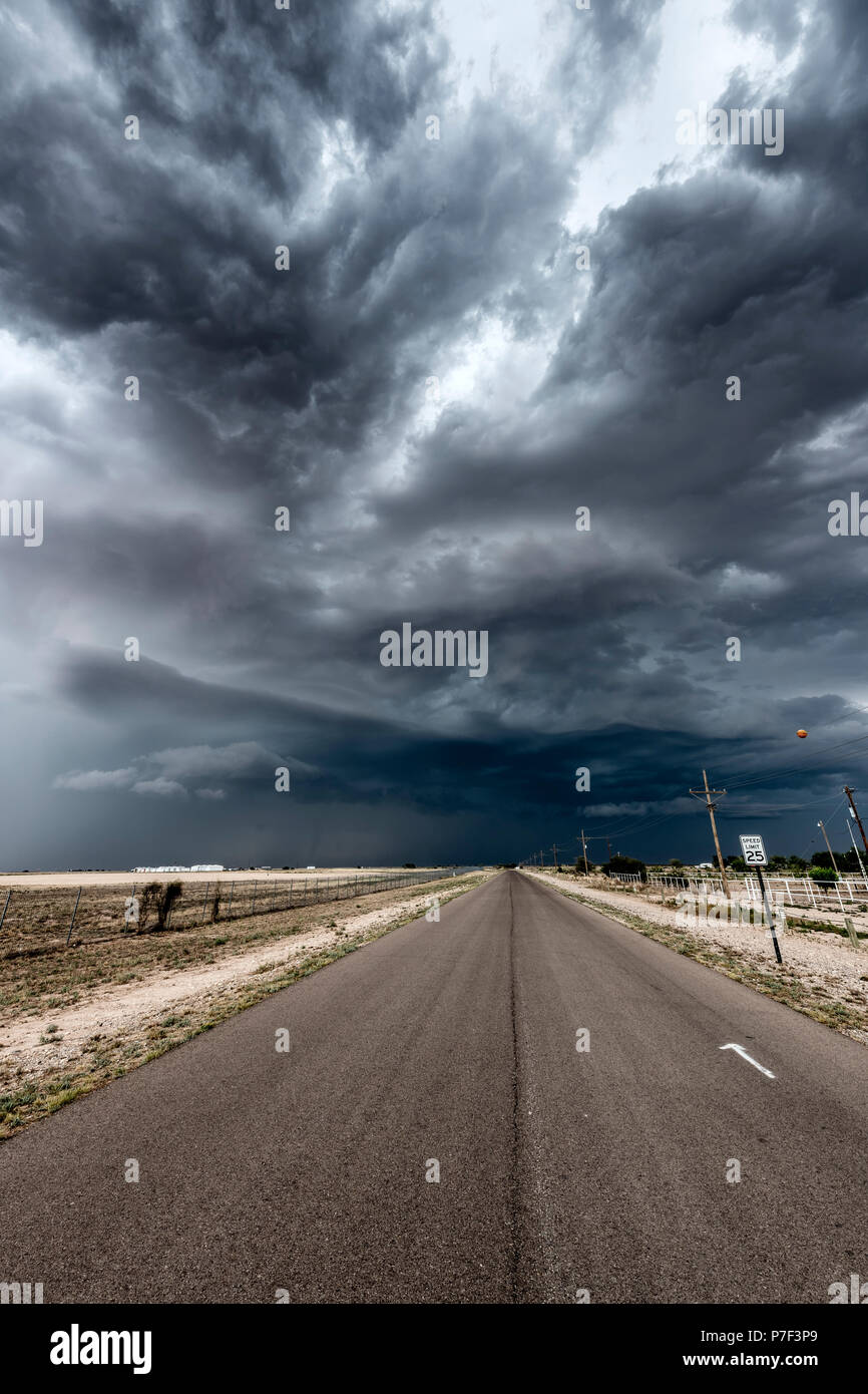 A massive, dark thunderstorm forming over a long, narrow road in