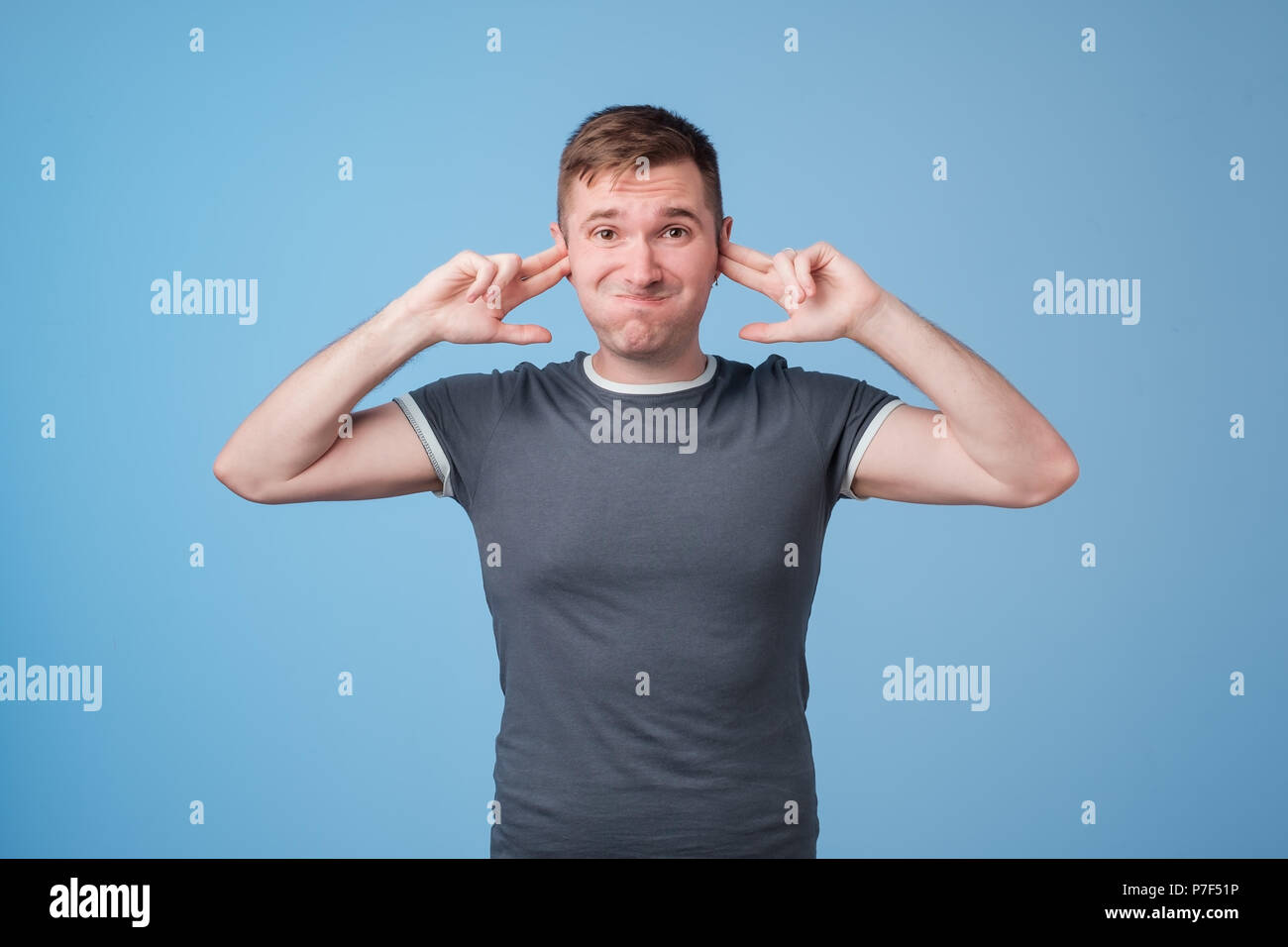 Studio portrait of young, unhappy, stressed man covering his ears, standing on blue wall background. Stock Photo