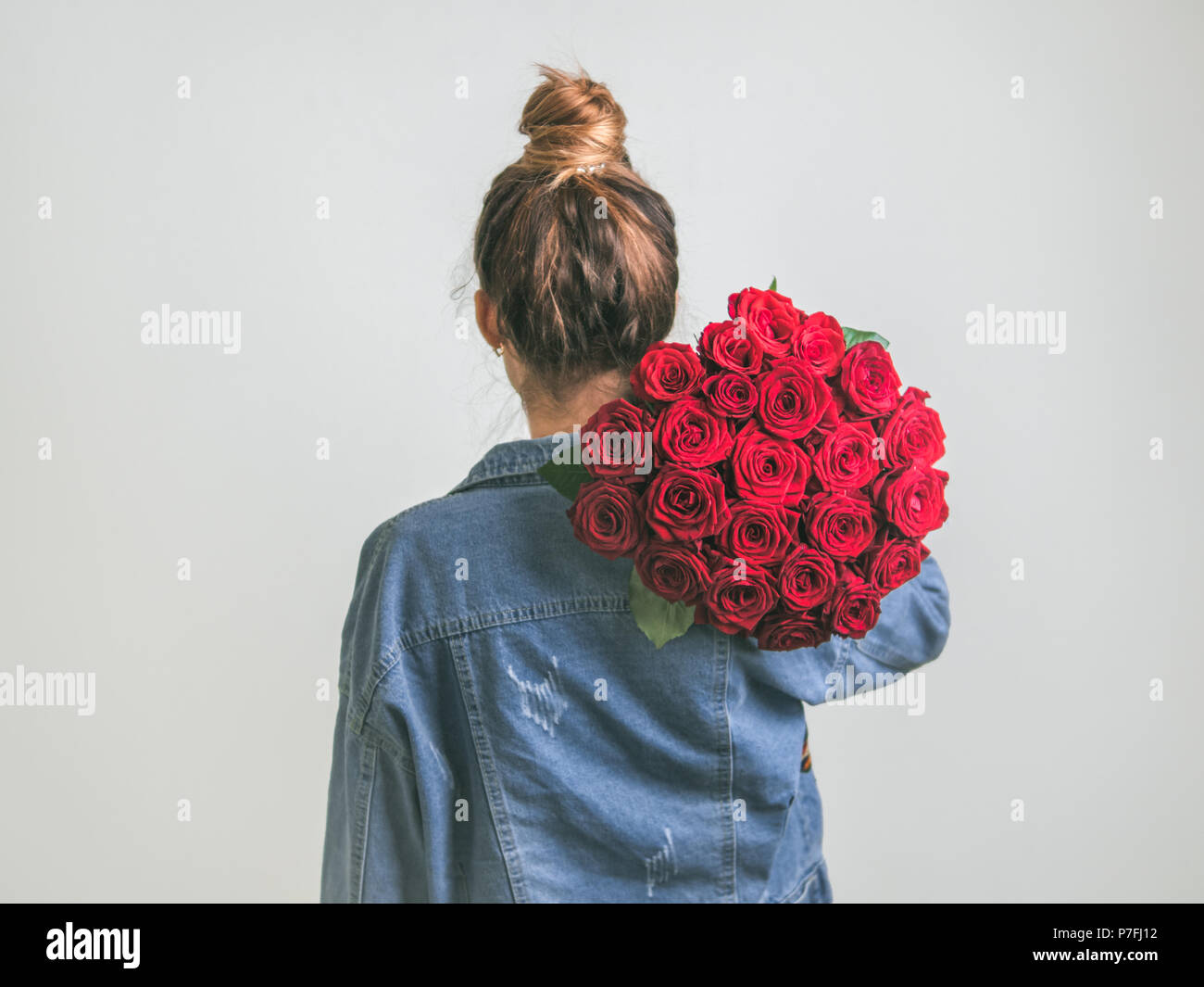 Back view of young woman in denim jacket holding bunch of red roses on shoulder. Girl with bun updo in jeans holding flowers. White background.Copy sp Stock Photo