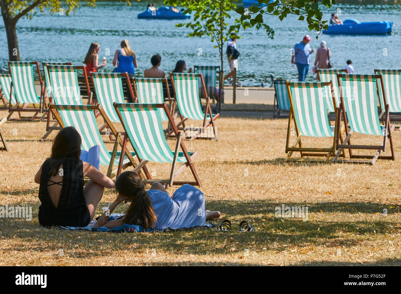 Two young women sitting in the sunshine in Hyde Park, London UK, during the July 2018 heatwave, with deck chairs and boats on the lake, in  the background Stock Photo