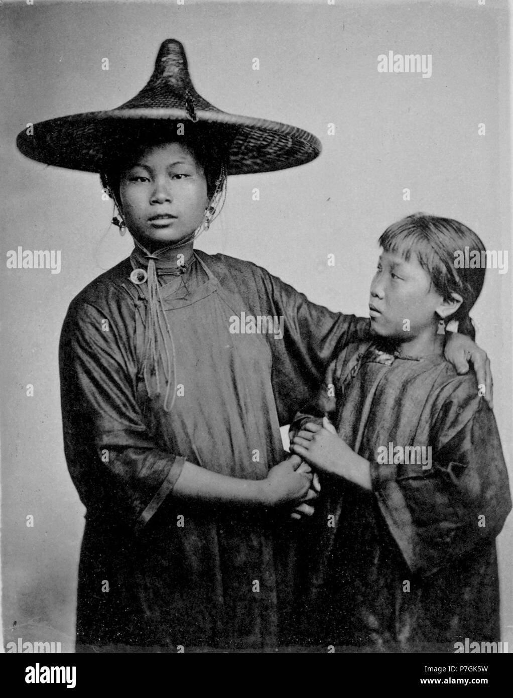English: John Thomson: THESE are the two daughters of a respectable boating family. They have been trained to the use of the oar, and the management of boats, from earliest childhood. Happy for them they are not slaves, purchased by some designing dame, and destined for a worse fate than the life of careful industry common to the labouring poor of Canton. The hat worn by the elder sister is made of ratan, closely woven, and varnished so as to render it waterproof; it affords protection from the sun as well as the rain, and serves, indeed, all the purposes of an umbrella. It has, too, this adva Stock Photo