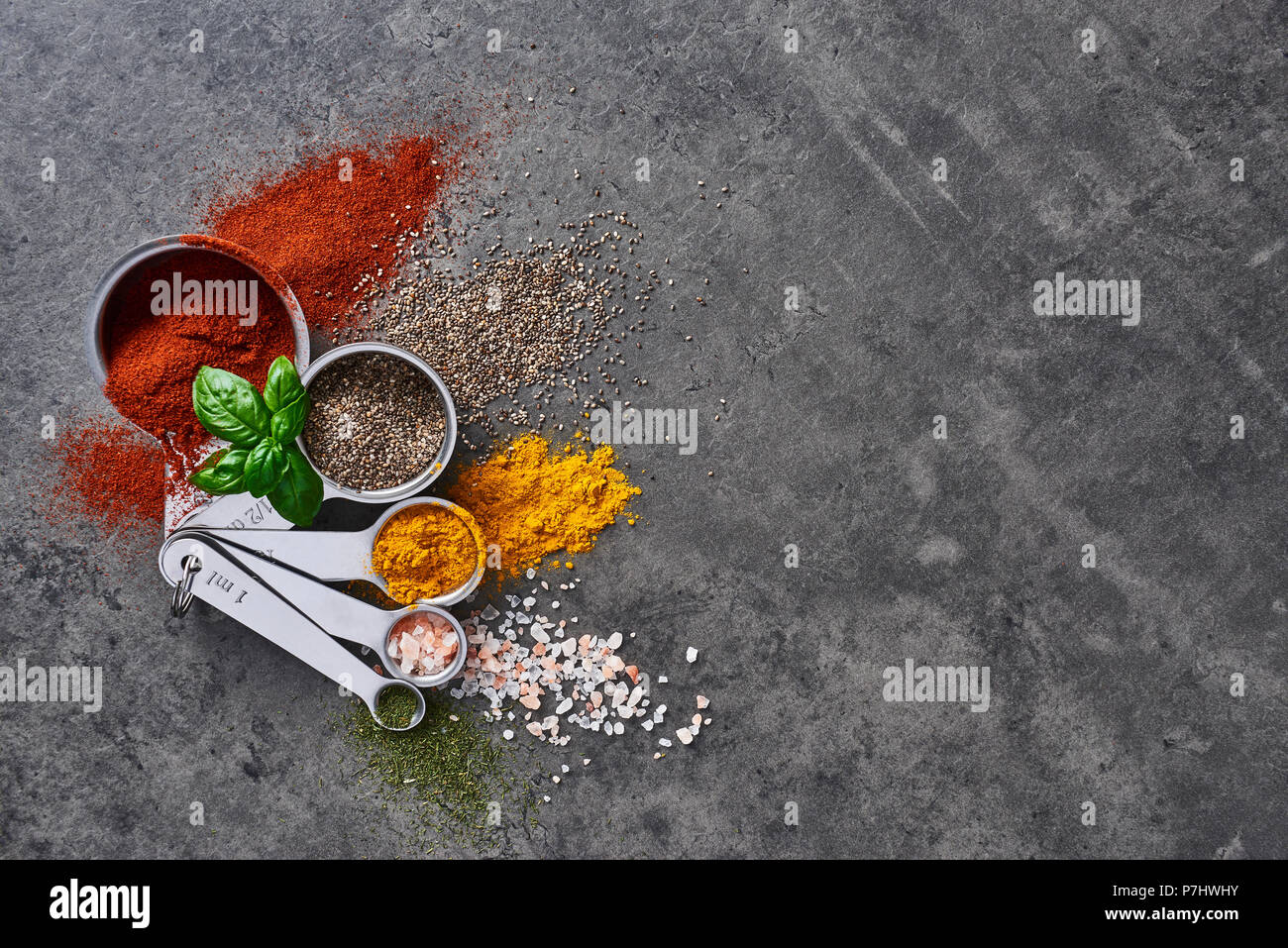 Flat lay of various spices in measuring cups with fresh basil on stone table. Top view with copy