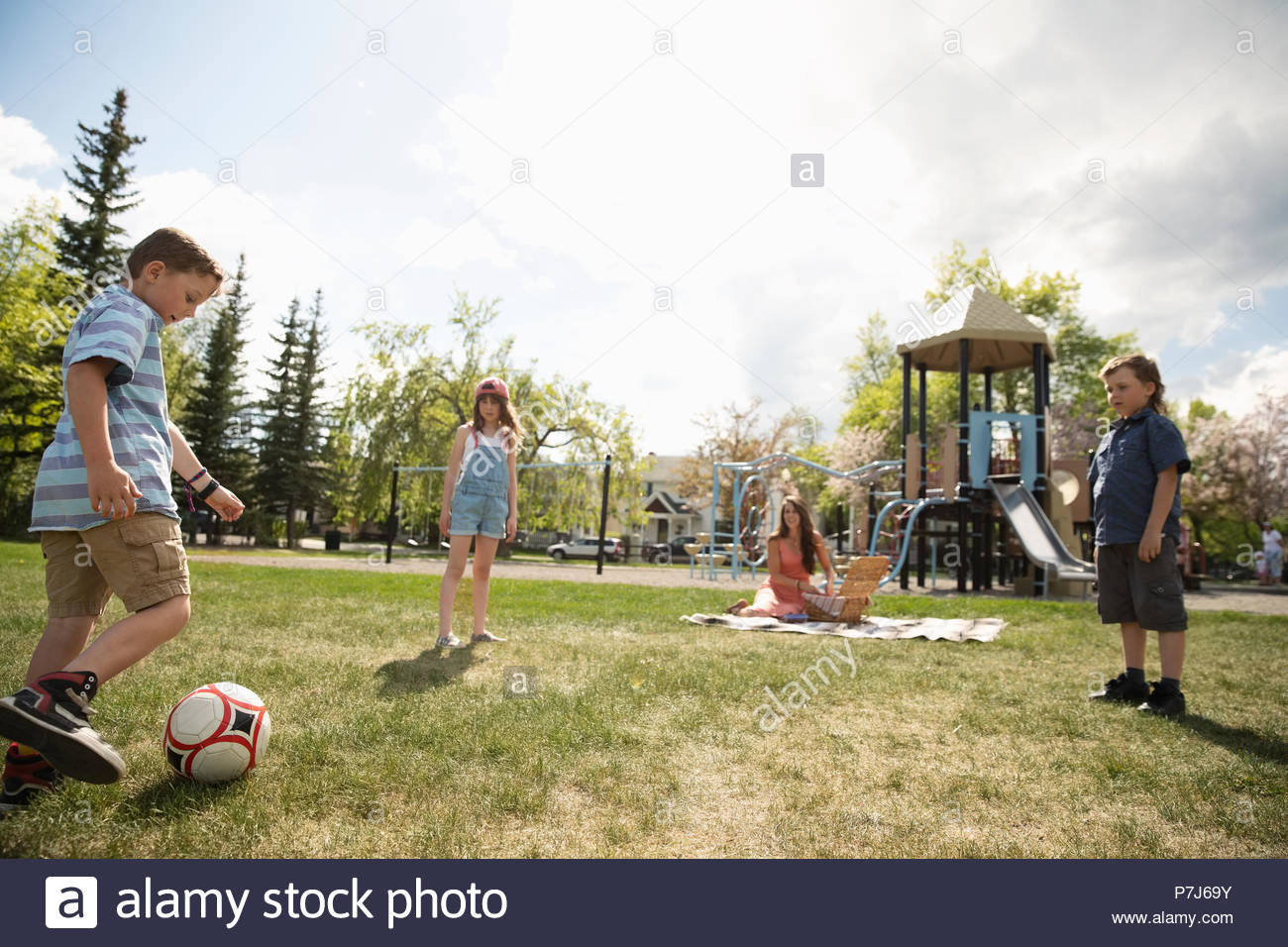 Mother on picnic blanket watching kids playing soccer in sunny park Stock Photo