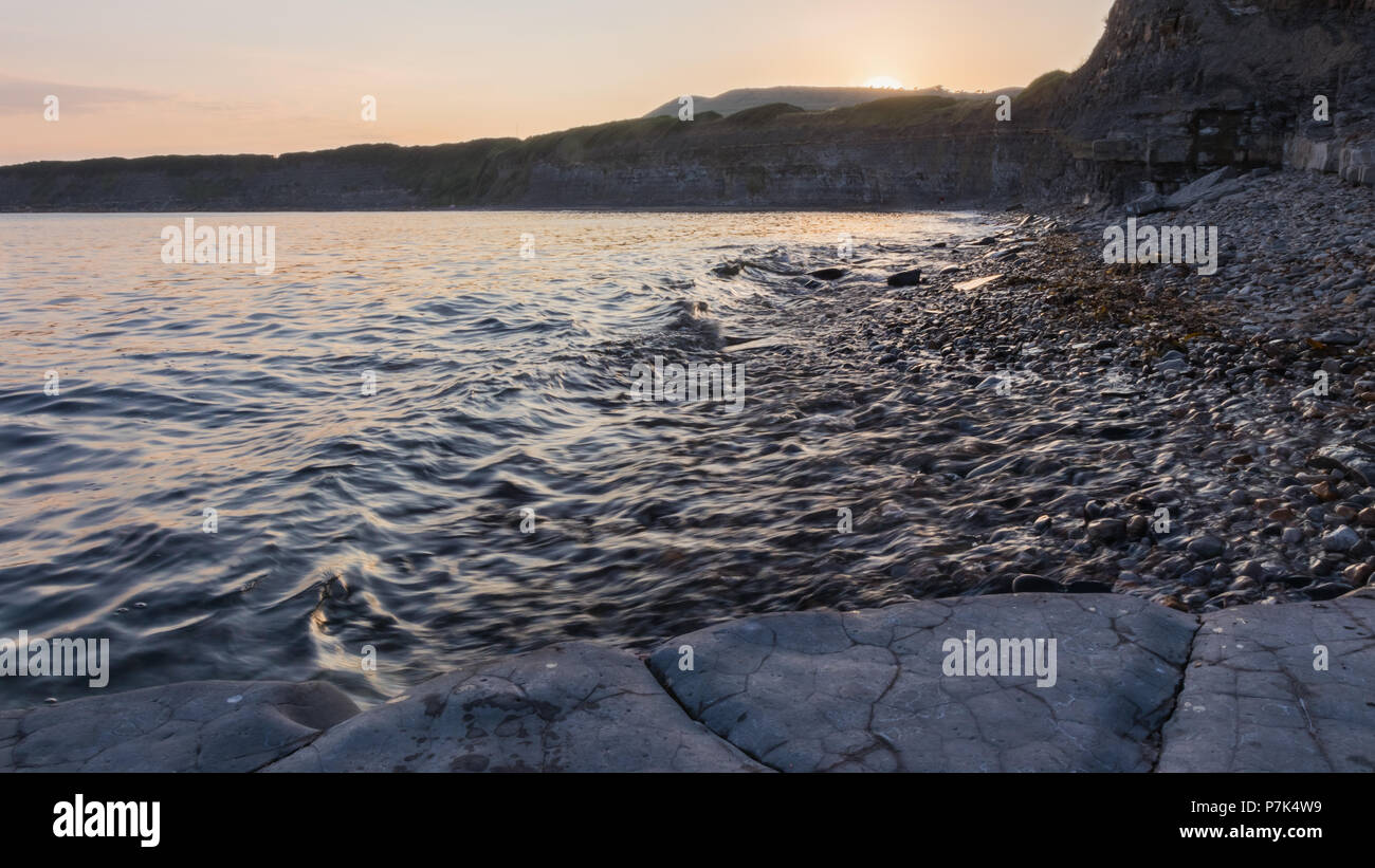 Kimmeridge bay cliffs in Dorset, UK as seen from sea level during a quiet summer sunset with small crashing waves and the setting sun. Stock Photo