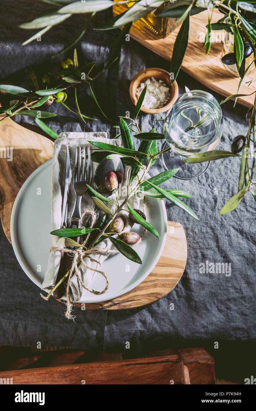 Table setting with gray Linen tablecloth and napkin, white plate, cutlery and and olive tree branch boards decoration. Top view. Stock Photo