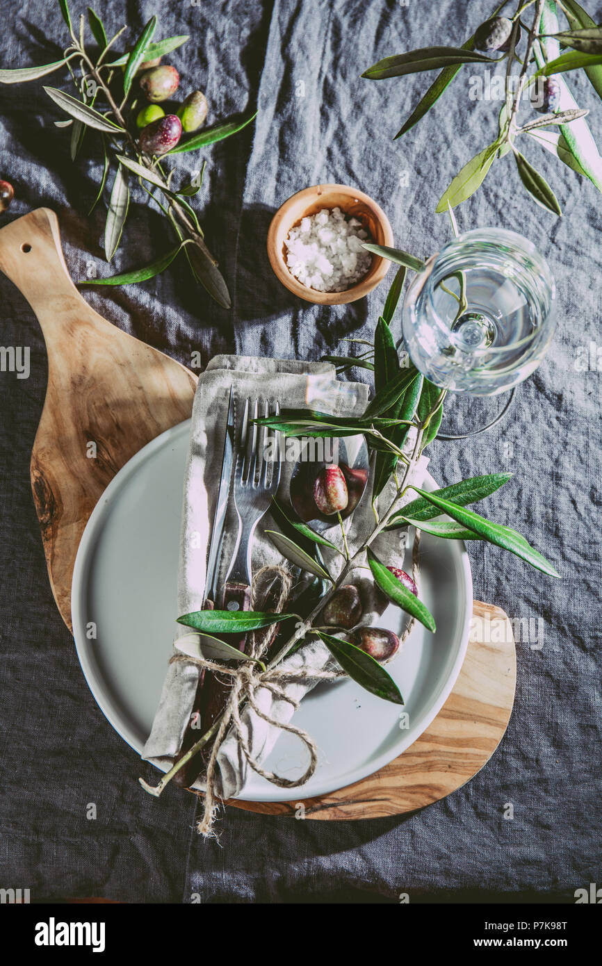 Table setting with gray Linen tablecloth and napkin, white plate, cutlery and and olive tree branch boards decoration. Top view. Stock Photo