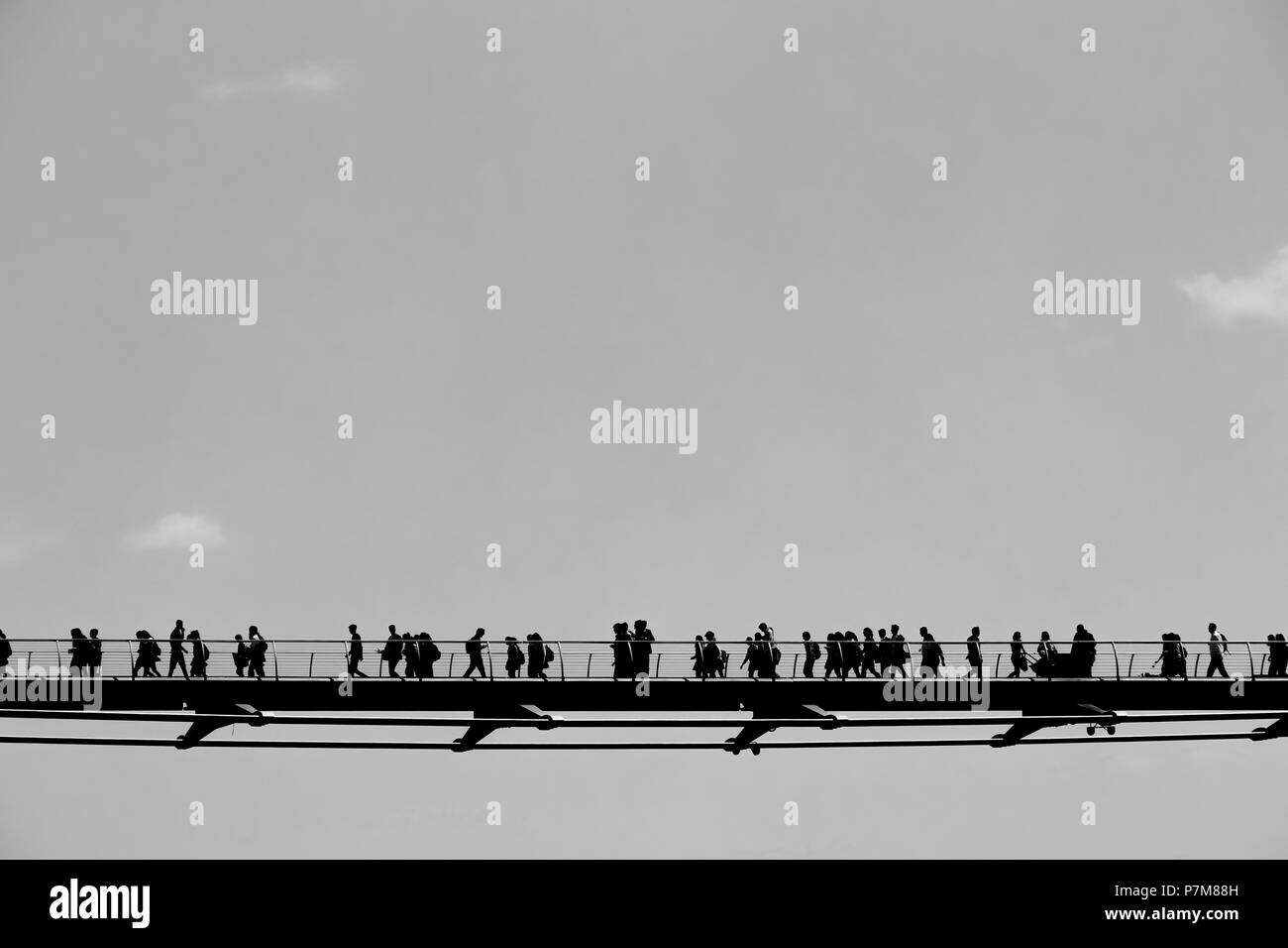 Many pedestrians walking across the London Millennium Footbridge are silhouetted against sky that has 3 little white clouds; copy space, black & white. Stock Photo